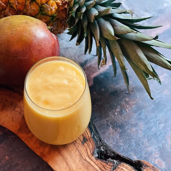 A glass of yellow smoothie sits on a wooden board next to a mango and a pineapple.