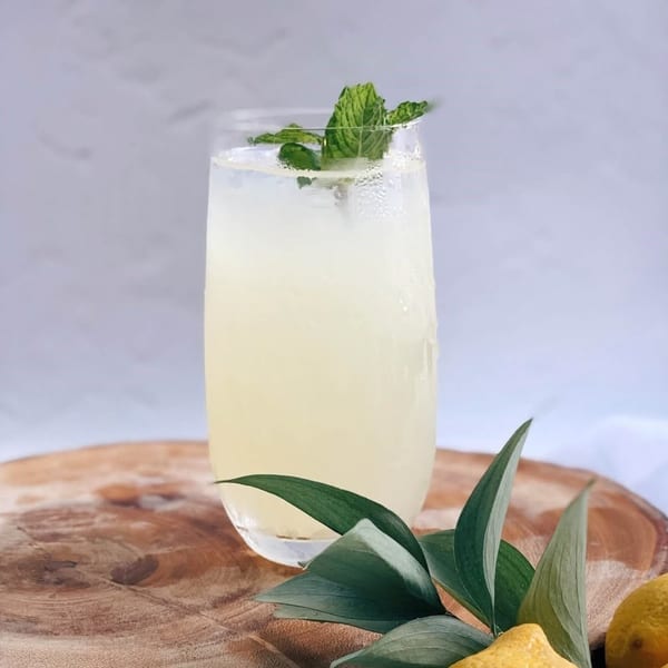 A refreshing glass of lemonade with mint garnish, lemons, and green leaves on a wooden board.